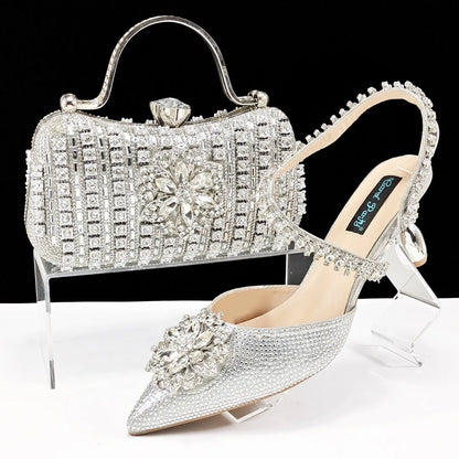 Women's black high-heel ankle strap shoes with floral rhinestone buckle and matching clutch bag on a neutral background