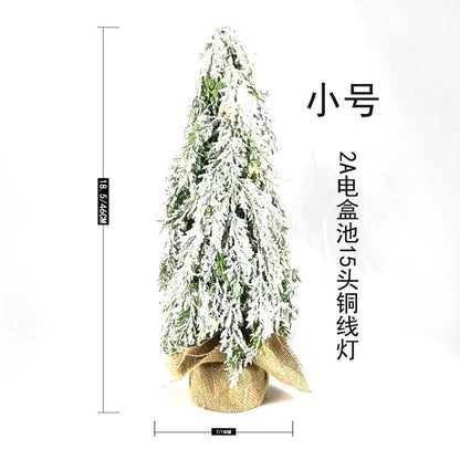 Small white artificial Christmas tree on a table with LED lights and a snow sprayer unit at its base.