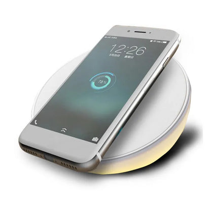 White wireless charging pad with digital clock display, alarm, and LED night light on a bedside table.