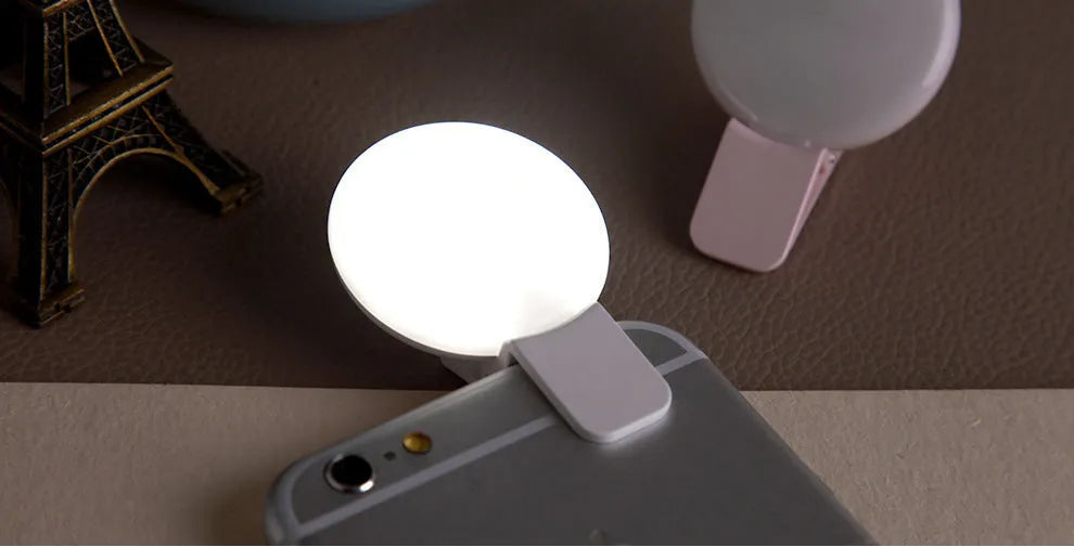 Round black mobile phone fill light with adjustable clip attached to a smartphone.