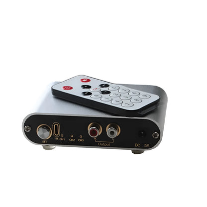 Silver aluminium audio source switch selector unit with infrared remote control on a black surface.