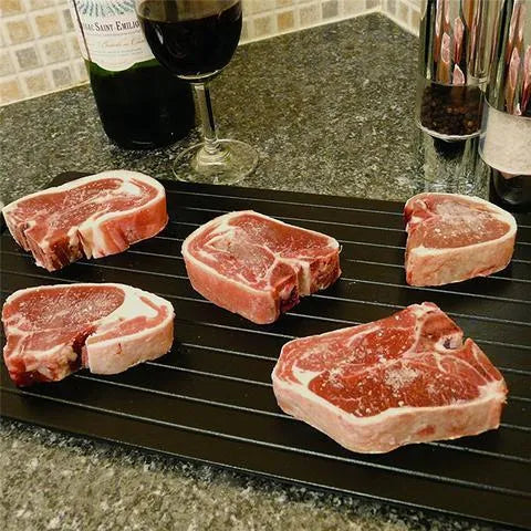 Black aluminium fast defrosting tray for kitchen use, shown thawing frozen meat.