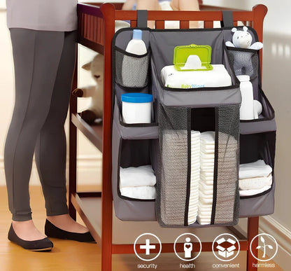 Grey baby nappy organiser attached to crib rail with Velcro straps, showing storage compartments for nappies and essentials