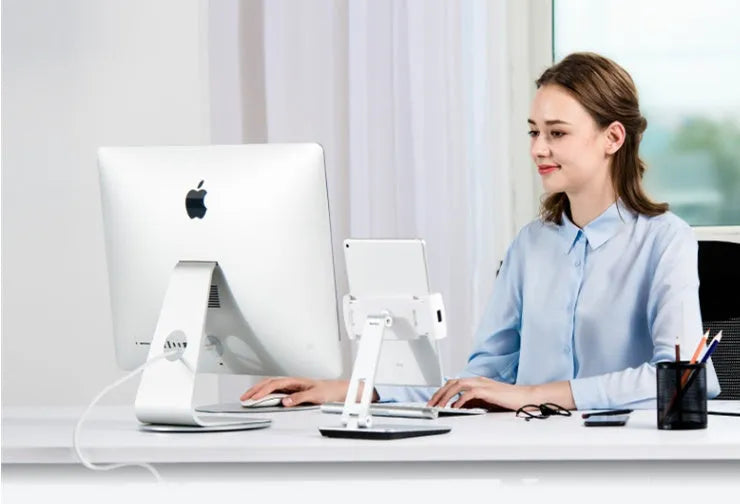 Silver foldable aluminium phone and tablet holder on a wooden desk, showing 360-degree rotation and silicone pads.