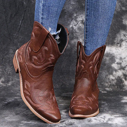 Women's brown embroidered ankle cowboy boots with a low wedge heel and pointed toe on a wooden floor.