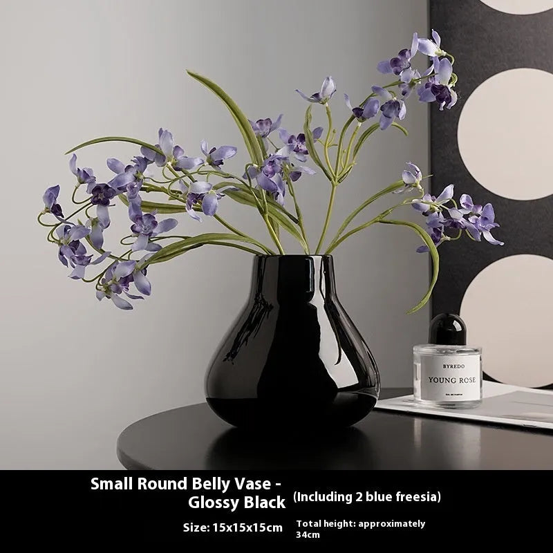 Black ceramic vase with round belly design and two white artificial orchid stems on wooden table