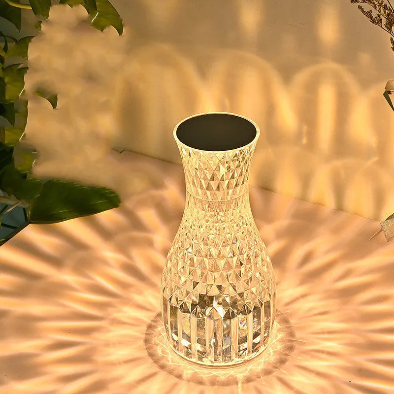 Transparent vase-shaped crystal table lamp with remote control on a wooden bedside table.