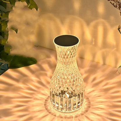 Transparent vase-shaped crystal table lamp with remote control on a wooden bedside table.