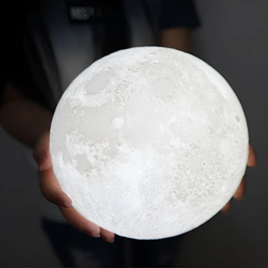 3D printed white moon lamp glowing on a wooden stand, showing detailed crater texture on its surface.