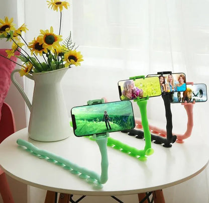 Black flexible gooseneck phone holder with suction cup mounted on a desk, holding a mobile phone at an angle.