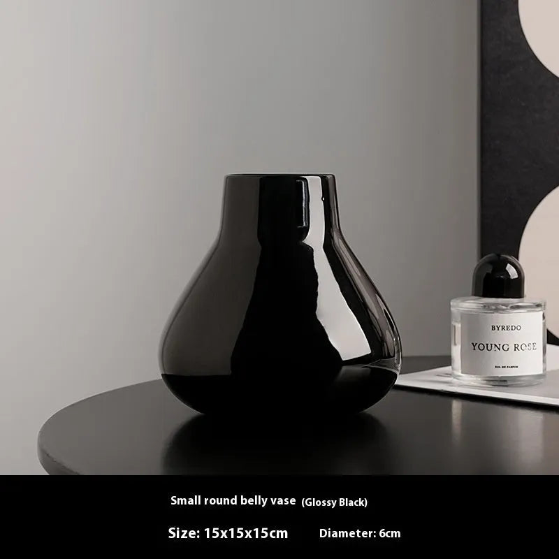 Black ceramic vase with round belly design and two white artificial orchid stems on wooden table