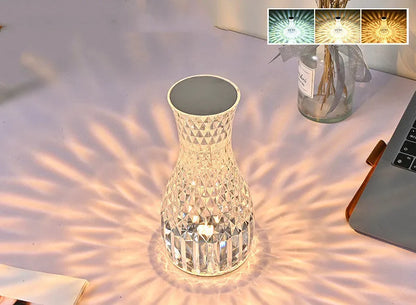 Transparent vase-shaped crystal table lamp with remote control on a wooden bedside table.
