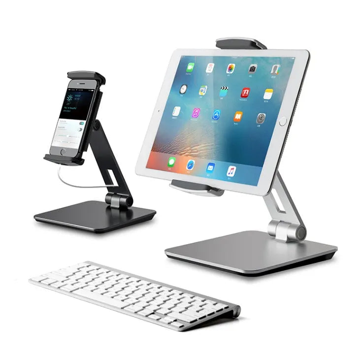 Silver foldable aluminium phone and tablet holder on a wooden desk, showing 360-degree rotation and silicone pads.