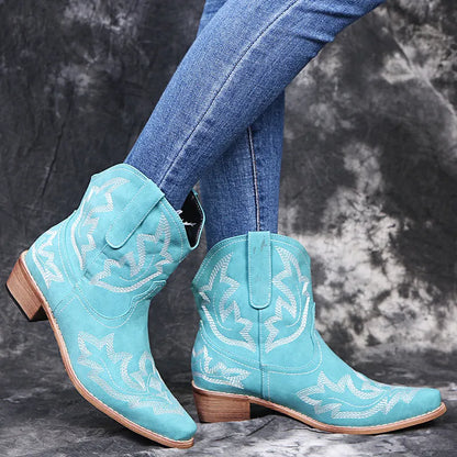 Women's blue embroidered cowboy ankle boots with a low wedge heel and pointed toe on a wooden floor.