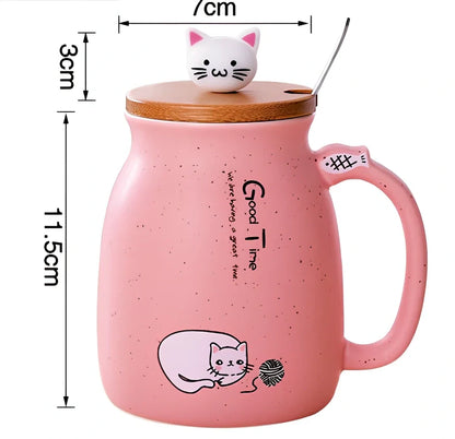 Pink ceramic mug with cartoon cat design, wooden lid, and stainless steel spoon on table