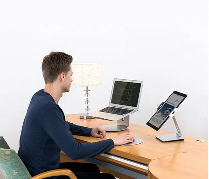 Silver foldable aluminium phone and tablet holder on a wooden desk, showing 360-degree rotation and silicone pads.