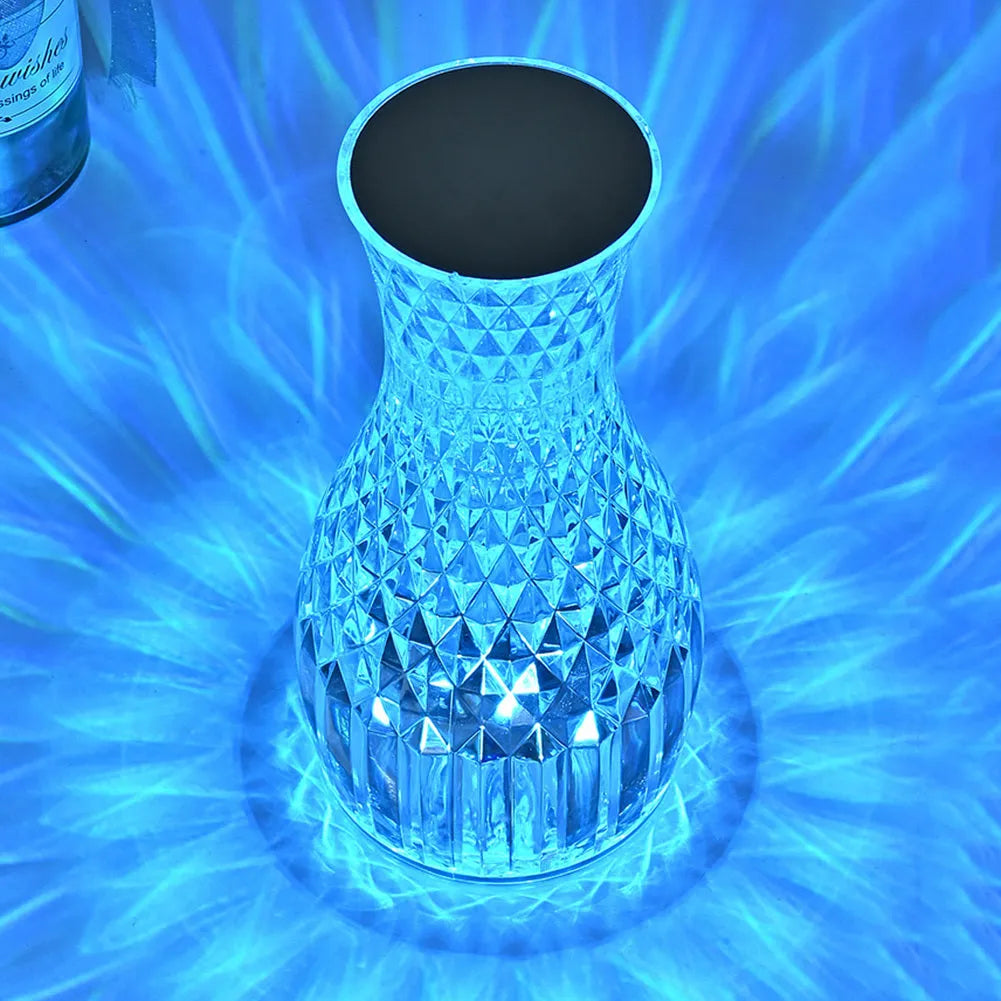 Transparent vase-shaped crystal table lamp with remote control on a wooden bedside table.