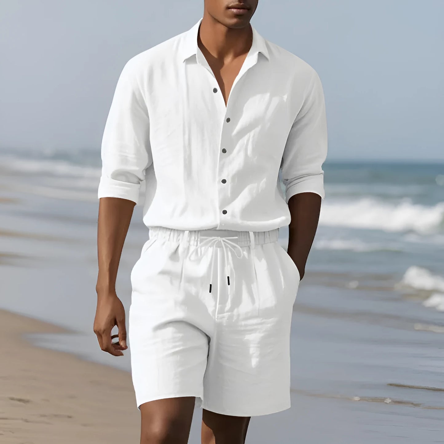 A man wearing a light blue linen long sleeve shirt and matching shorts on the beach.