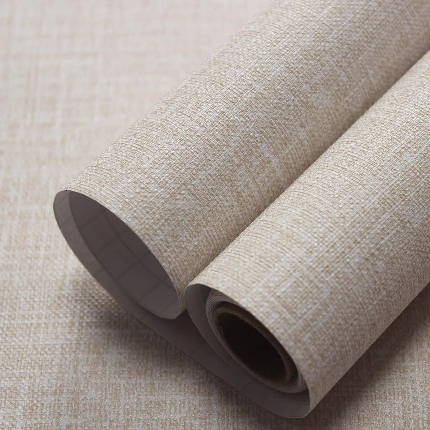 Roll of beige linen self-adhesive wallpaper with textured finish next to a sample applied to a clean white wall.