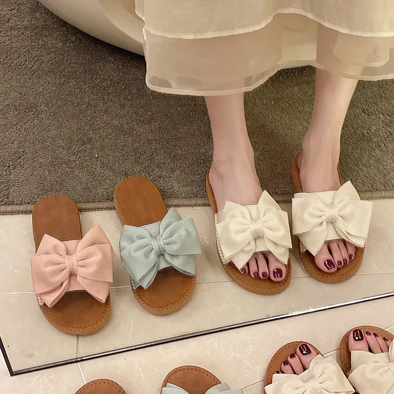 Women's beige suede slip-on sandals with a flat rubber sole and decorative bow.