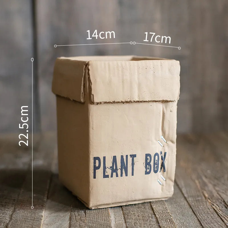 Square cement planter with matte finish and drainage holes, next to a small green plant.