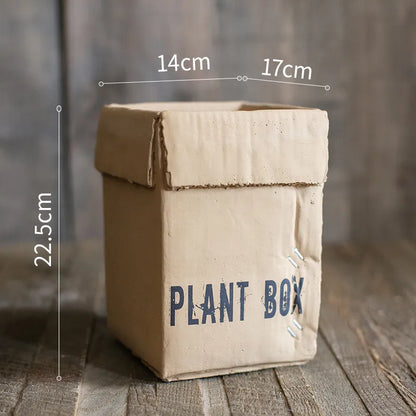 Square cement planter with matte finish and drainage holes, next to a small green plant.