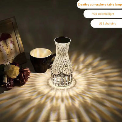 Transparent vase-shaped crystal table lamp with remote control on a wooden bedside table.