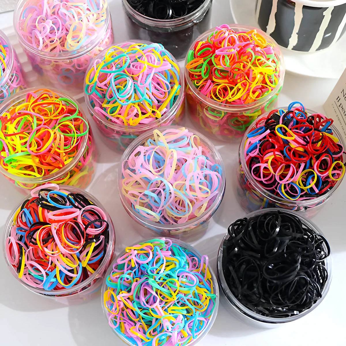 Multicoloured assortment of thin TPU elastic hair bands for babies and children in a reusable plastic container.
