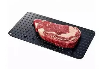 Black aluminium fast defrosting tray for kitchen use, shown thawing frozen meat.