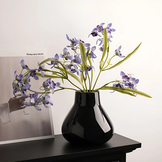 Black ceramic vase with round belly design and two white artificial orchid stems on wooden table