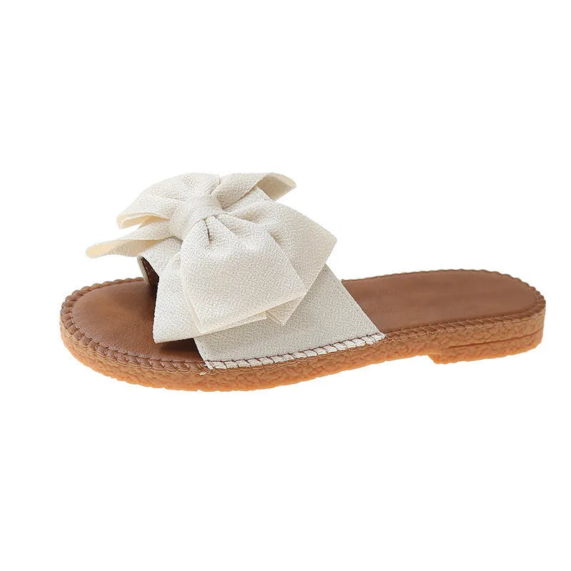 Women's beige suede slip-on sandals with a flat rubber sole and decorative bow.