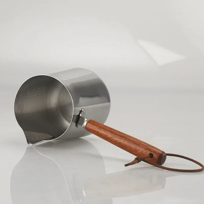 Stainless steel barista milk jug with wooden handle and internal measurement scale for latte art.
