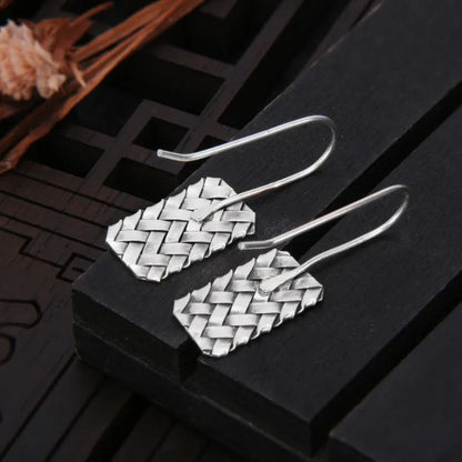 Close-up of a pair of silver geometric stud earrings with a woven diamond plaid pattern on a white background.