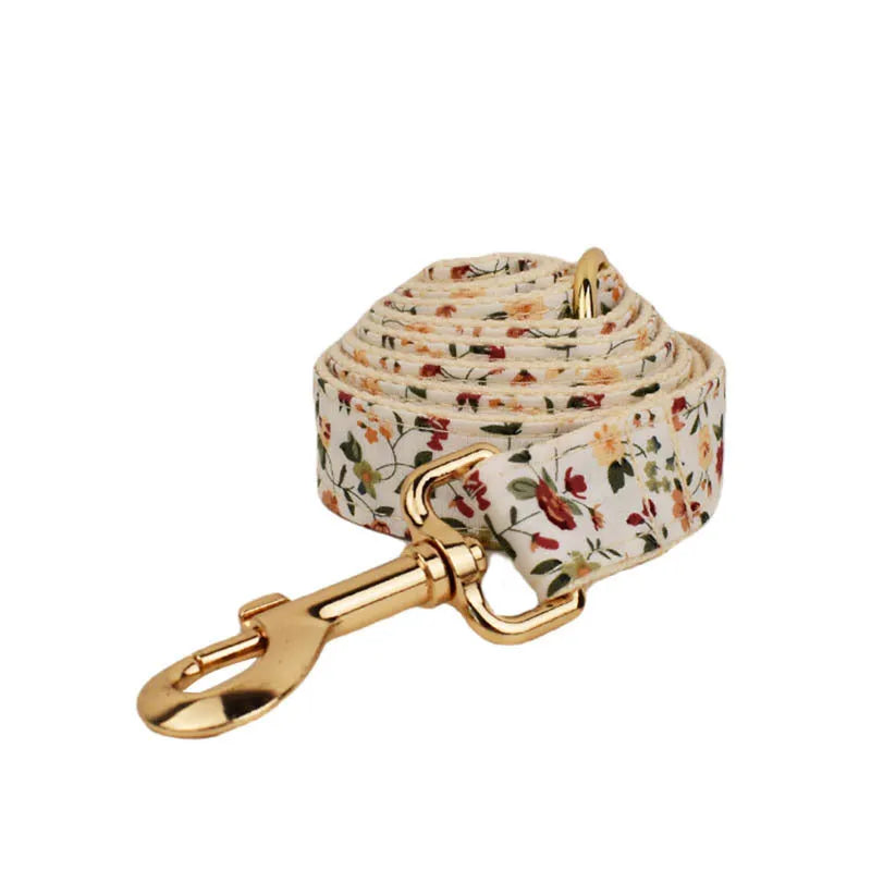 Small white flower patterned dog collar, leash, vest, bow, and poop bag holder set laid out flat.