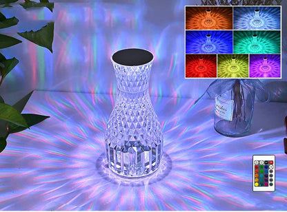 Transparent vase-shaped crystal table lamp with remote control on a wooden bedside table.