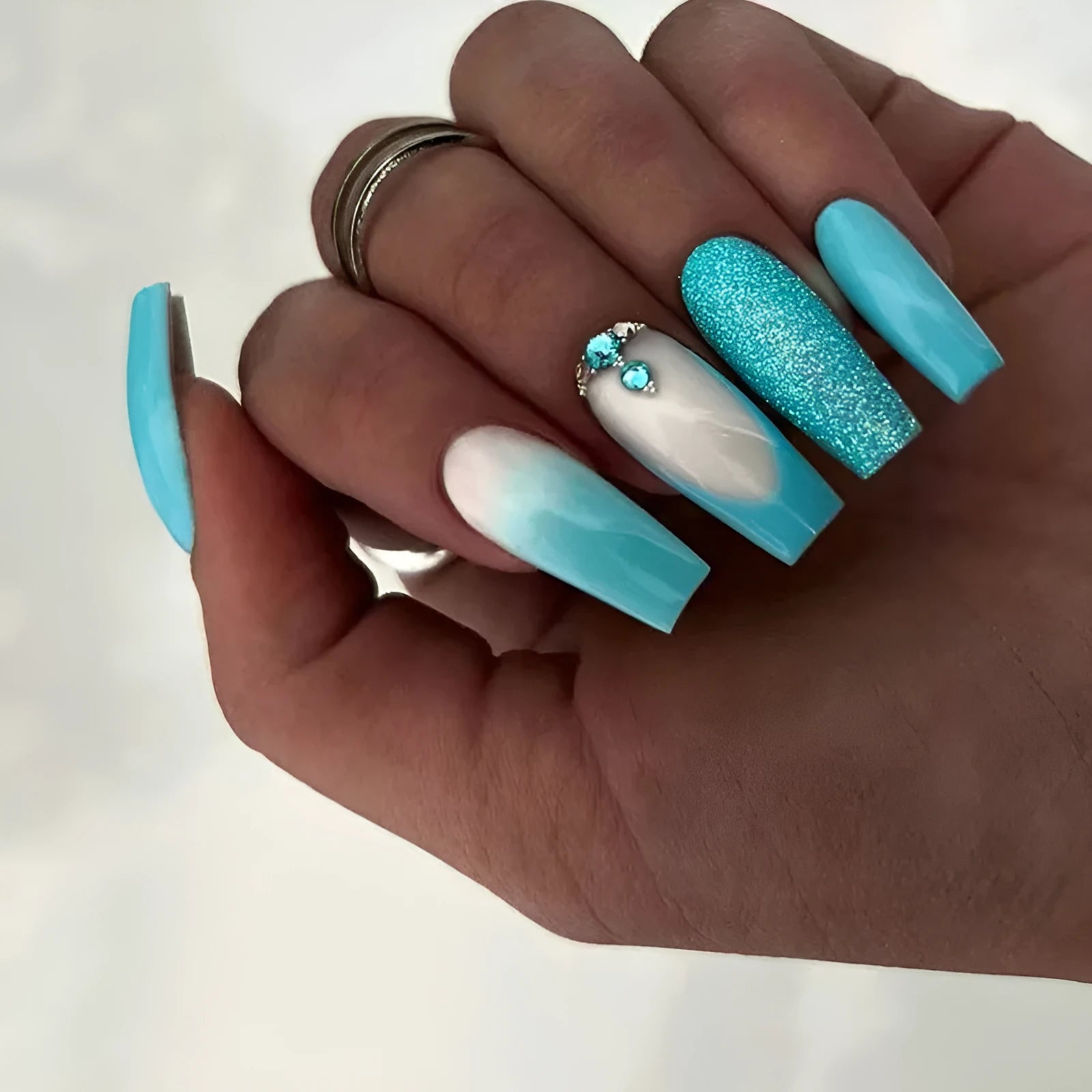 Set of long ballet-shaped false nails with smart blue colour and embedded diamond accents, next to a tube of adhesive glue.