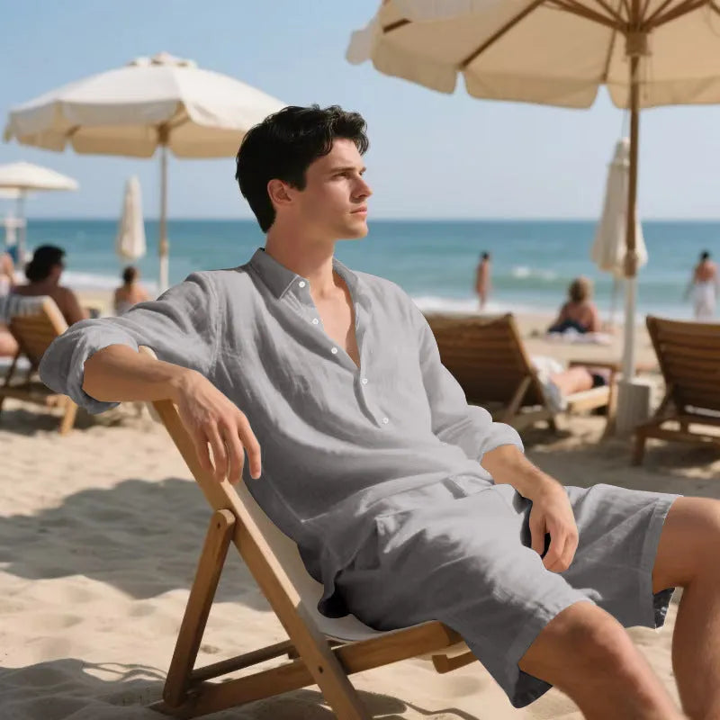 A man wearing a light blue linen long sleeve shirt and matching shorts on the beach.