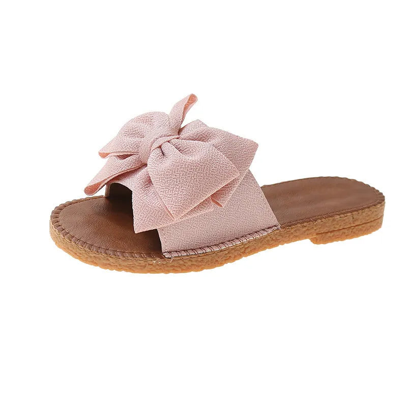 Women's beige suede slip-on sandals with a flat rubber sole and decorative bow.
