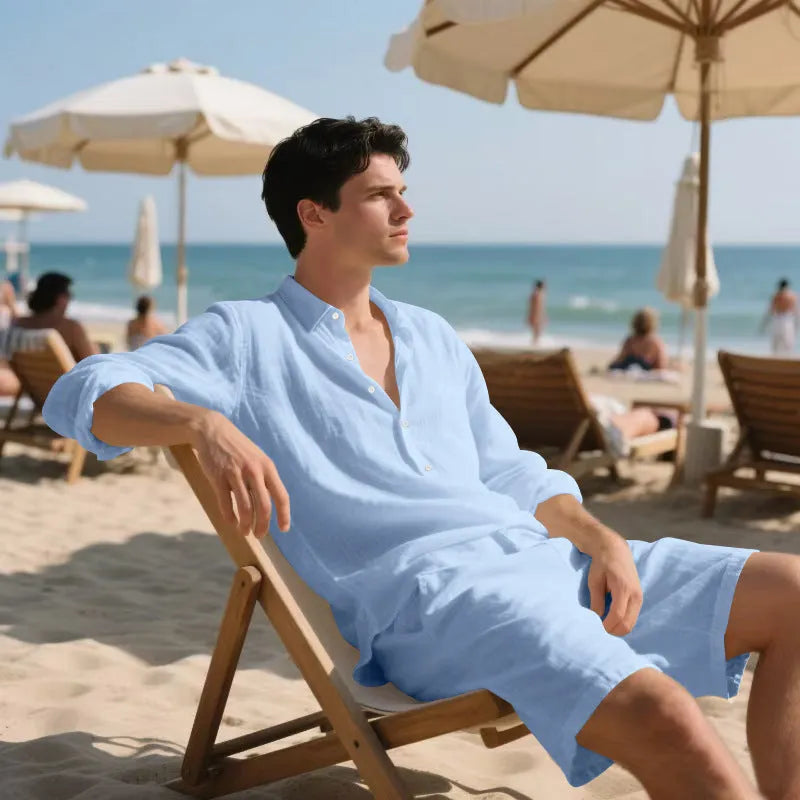 A man wearing a light blue linen long sleeve shirt and matching shorts on the beach.