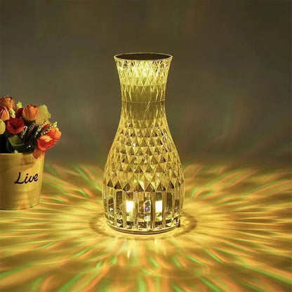Transparent vase-shaped crystal table lamp with remote control on a wooden bedside table.