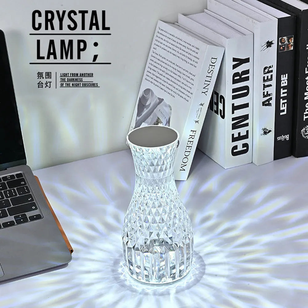 Transparent vase-shaped crystal table lamp with remote control on a wooden bedside table.