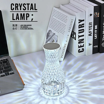 Transparent vase-shaped crystal table lamp with remote control on a wooden bedside table.