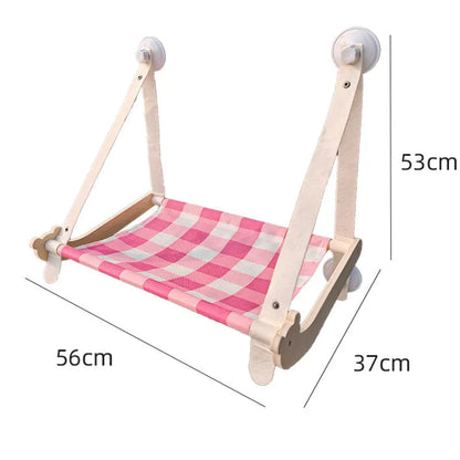 Pink cat hammock with wooden frame and suction cups attached to a window, with a cat resting inside.