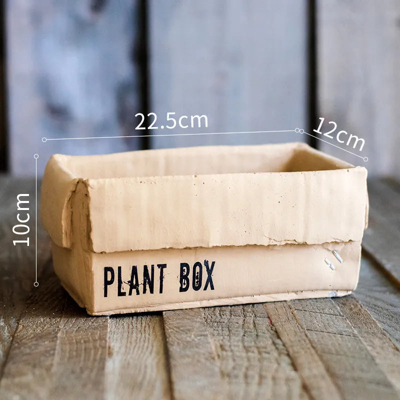 Square cement planter with matte finish and drainage holes, next to a small green plant.