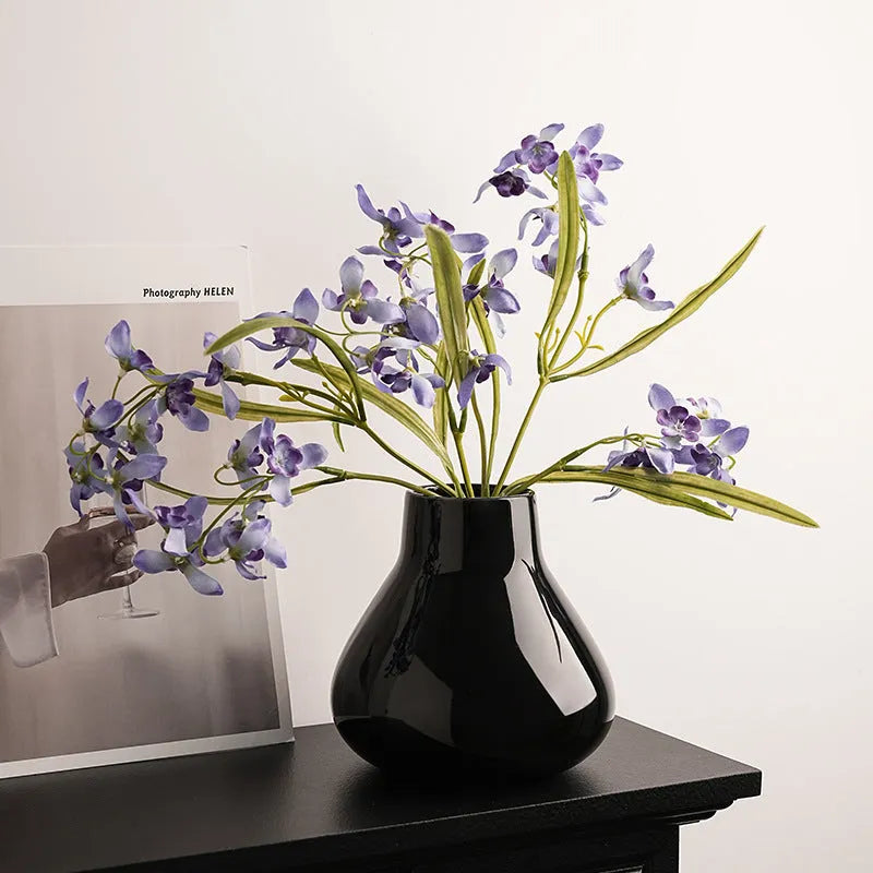 Black ceramic vase with round belly design and two white artificial orchid stems on wooden table