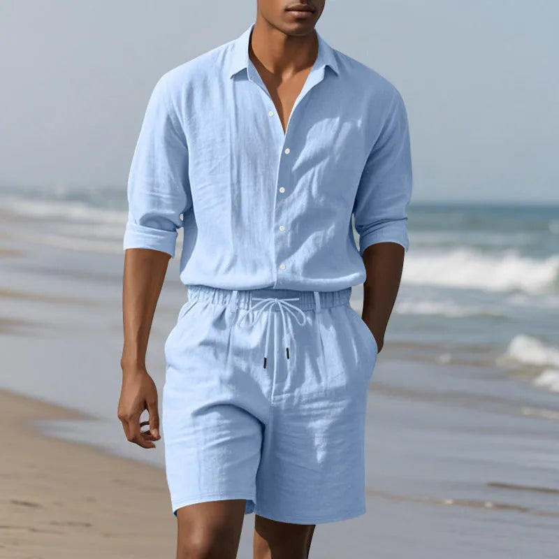 A man wearing a light blue linen long sleeve shirt and matching shorts on the beach.