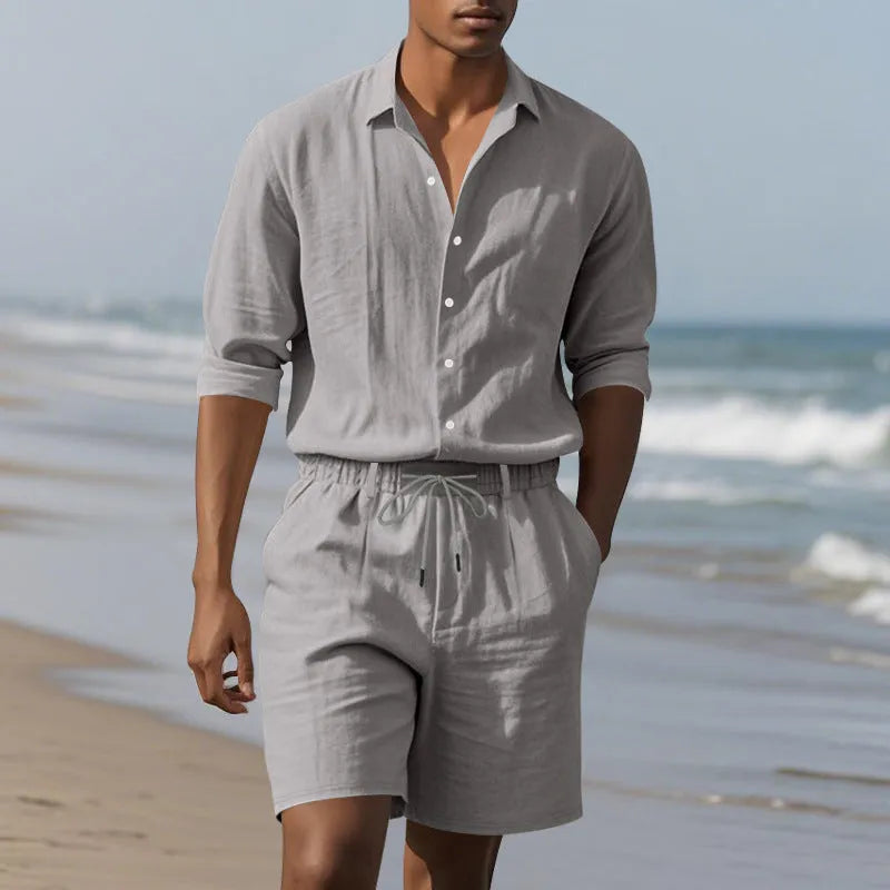 A man wearing a light blue linen long sleeve shirt and matching shorts on the beach.