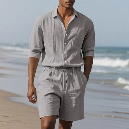 A man wearing a light blue linen long sleeve shirt and matching shorts on the beach.