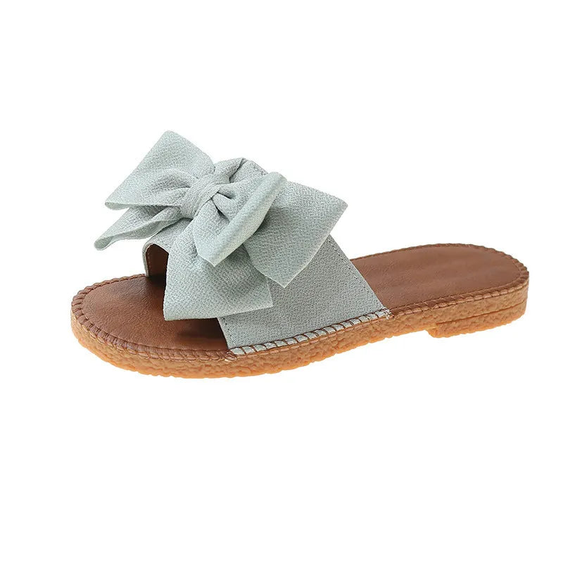 Women's beige suede slip-on sandals with a flat rubber sole and decorative bow.