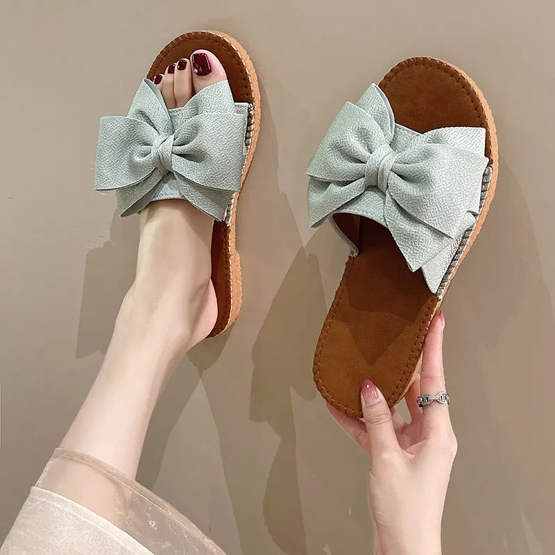 Women's beige suede slip-on sandals with a flat rubber sole and decorative bow.